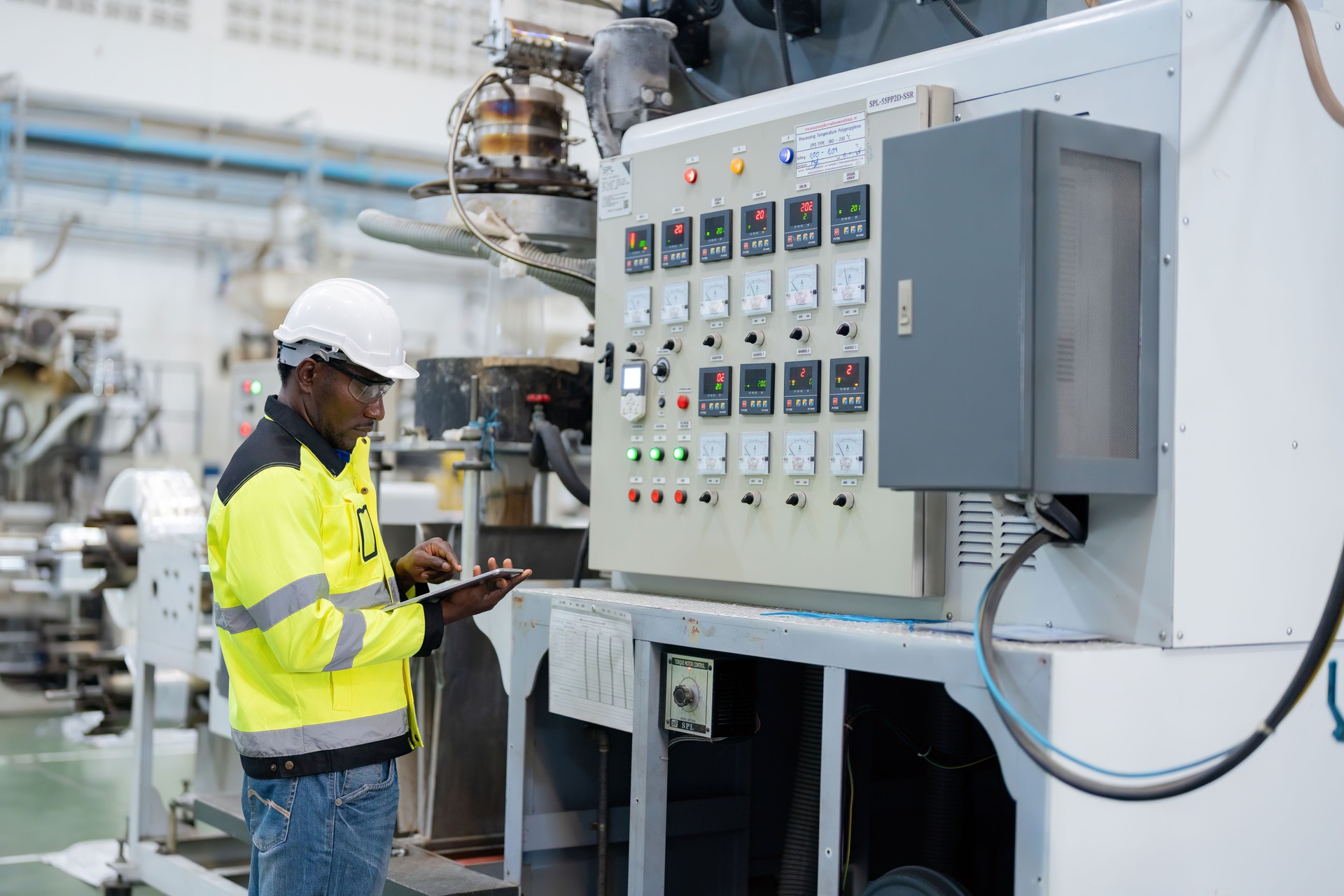 African male engineer Working and touching the tablet wear a helmet vest Standing near the machine, there is a Swiss button and a machine control board. In a large plastic and steel industry